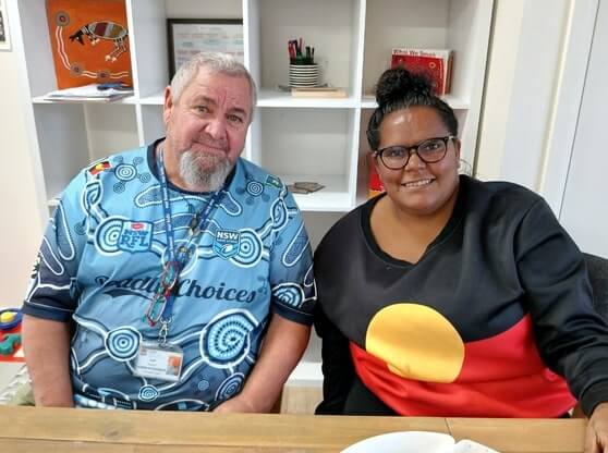 Two smiling individuals sit side by side at a table, wearing shirts that represent Aboriginal pride and health initiatives. One wears a blue shirt with circular patterns and logos including “NSW,” “Deadly Choices,” and the NRL. The other wears a black top featuring the Aboriginal flag.