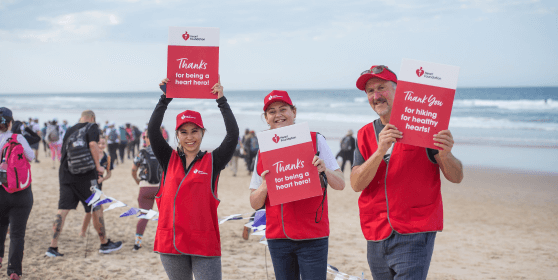 Three smiling volunteers in red vests stand on a beach holding signs that say “Thanks for being a heart hero” and “Thank you for hiking for healthy hearts.”