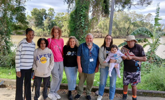Group of seven people, including one holding a baby, standing on a paved path beside a river with lush greenery and trees in the background.