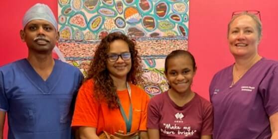 Two smiling individuals wear shirts representing Aboriginal pride and health initiatives, seated indoors with a shelf in the background.