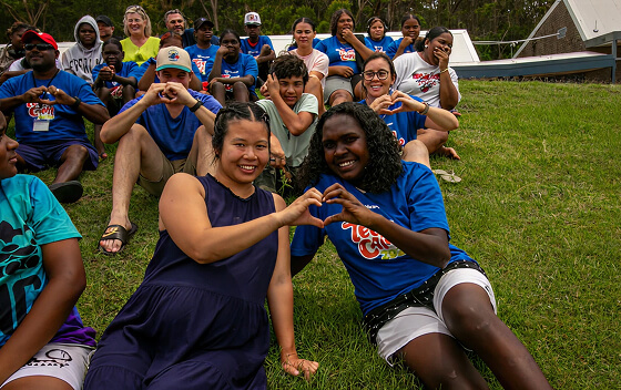 A diverse group of people sitting on grass making heart shapes with their hands.